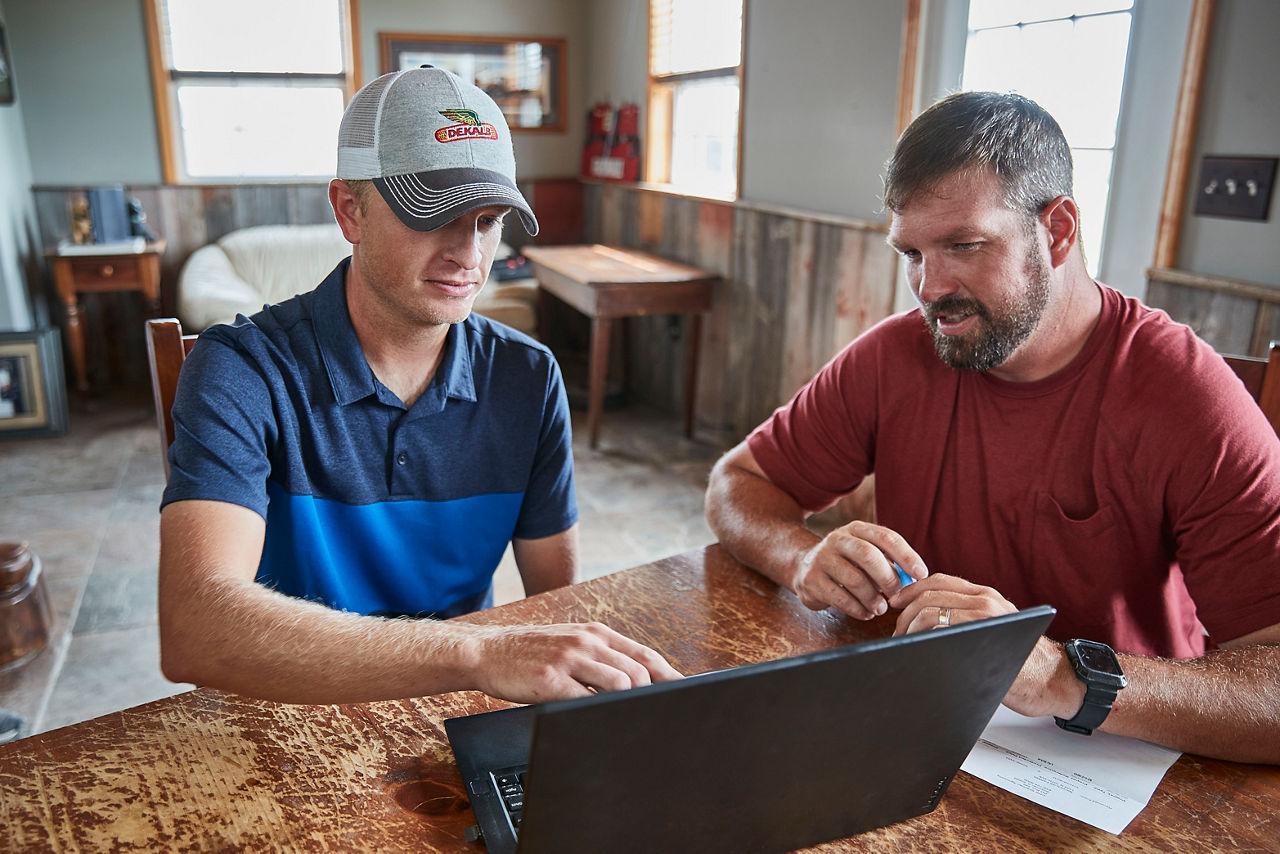 Two farmers discussing crop strategies while looking at a laptop, seated at a wooden table in a bright farmhouse. 